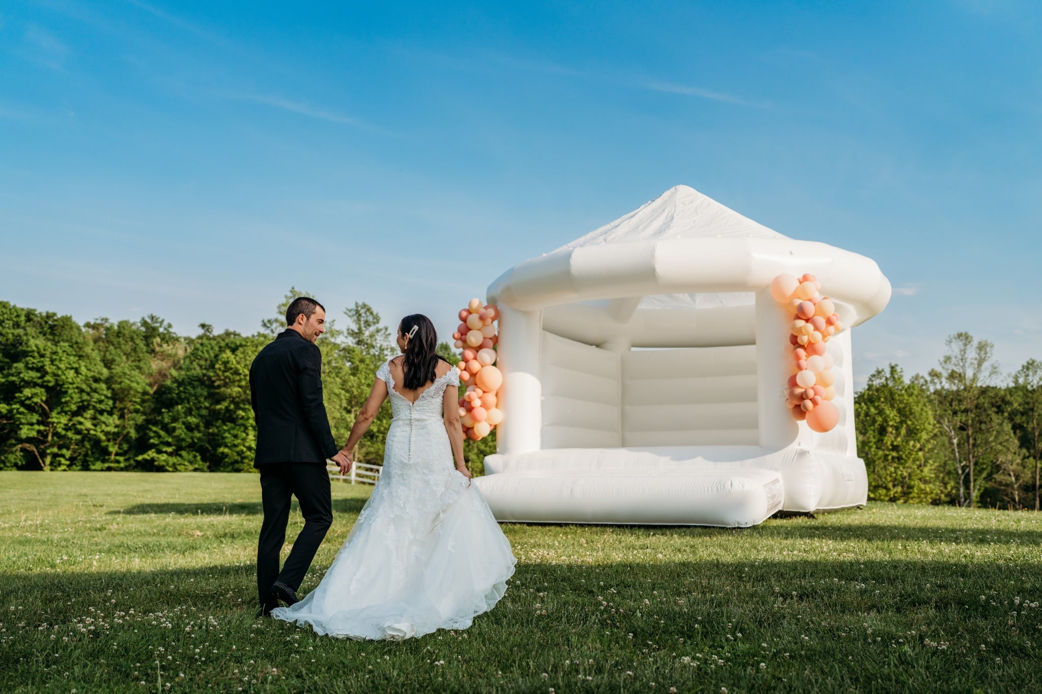 outdoor photo of a bride and groom walking towards a white castle bounce house